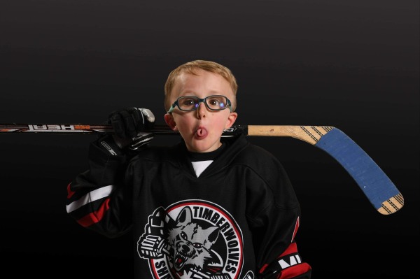 A photo featuring a young blonde boy making a taco tongue wearing, blue rimmed glasses a black, white and red hockey jersey and holding a stick across the back of his shoulders against a black background
